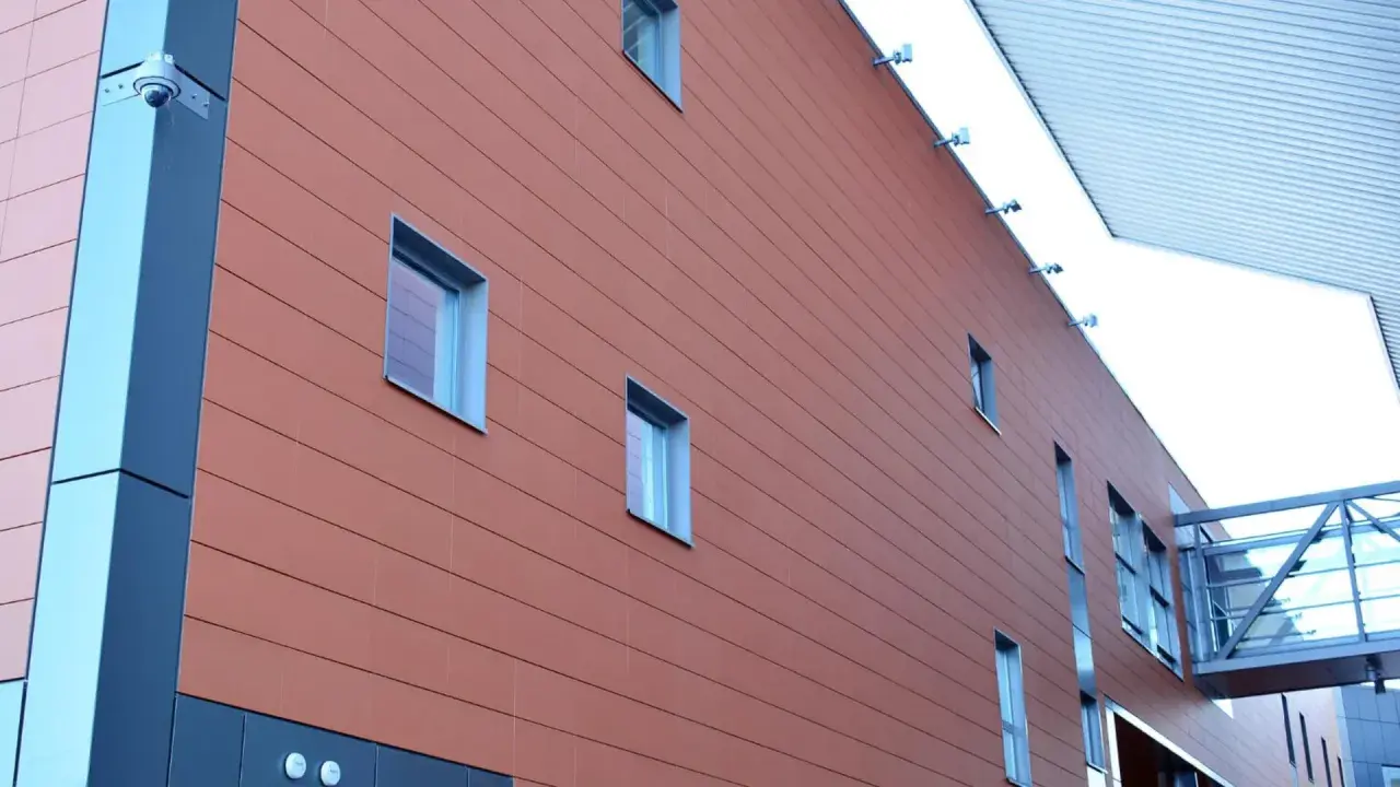Fachada moderna con revestimiento de sate en tono terracota, ventanas rectangulares y una pasarela acristalada.