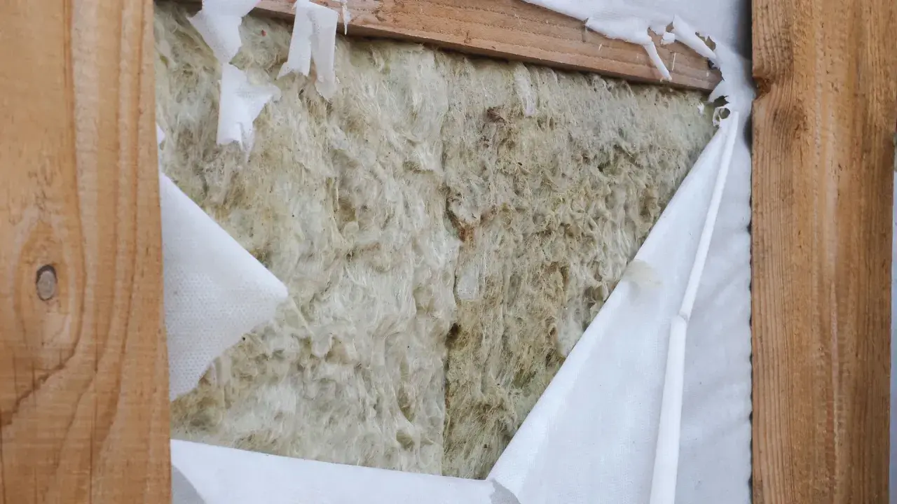 Aislamiento de lana de roca expuesto, con una barrera de vapor blanca rasgada que revela la textura fibrosa.