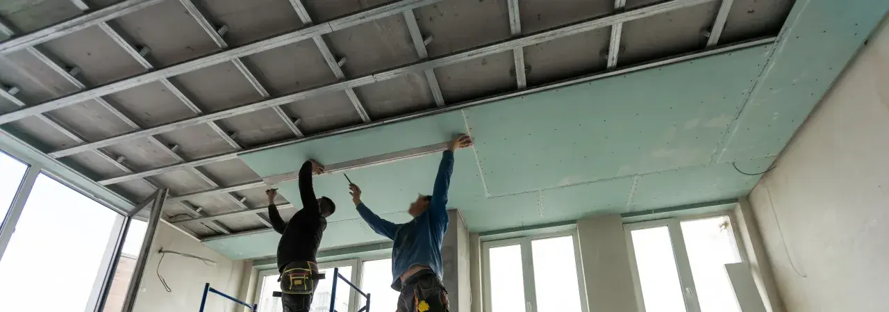 Dos trabajadores instalan placas de yeso para como aislar un techo por el interior. Se ve la estructura metálica del falso techo.