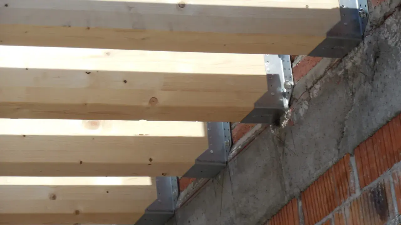 Detalle de la construcción: se están colocando vigas de madera sobre soportes metálicos anclados a una pared de ladrillo y hormigón.