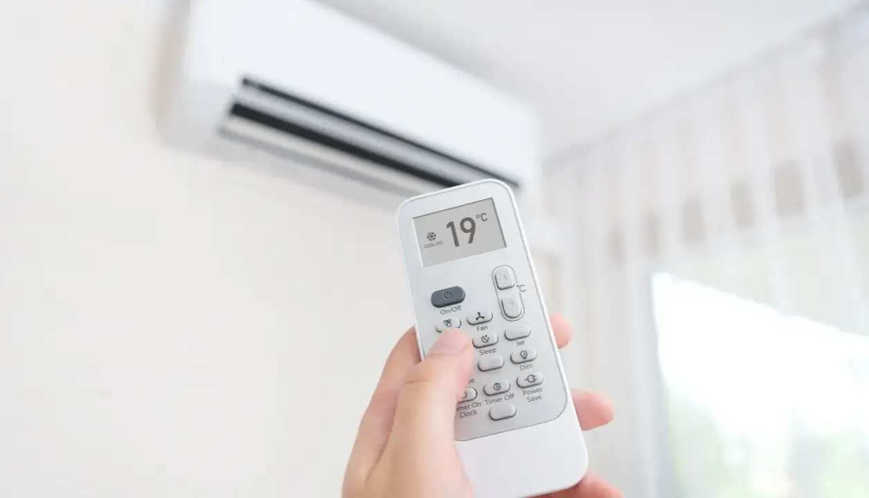 Mano sujetando control remoto de aire acondicionado, ajustando temperatura a 19°C. ¿Es mejor apagar el aire acondicionado o dejarlo encendido?