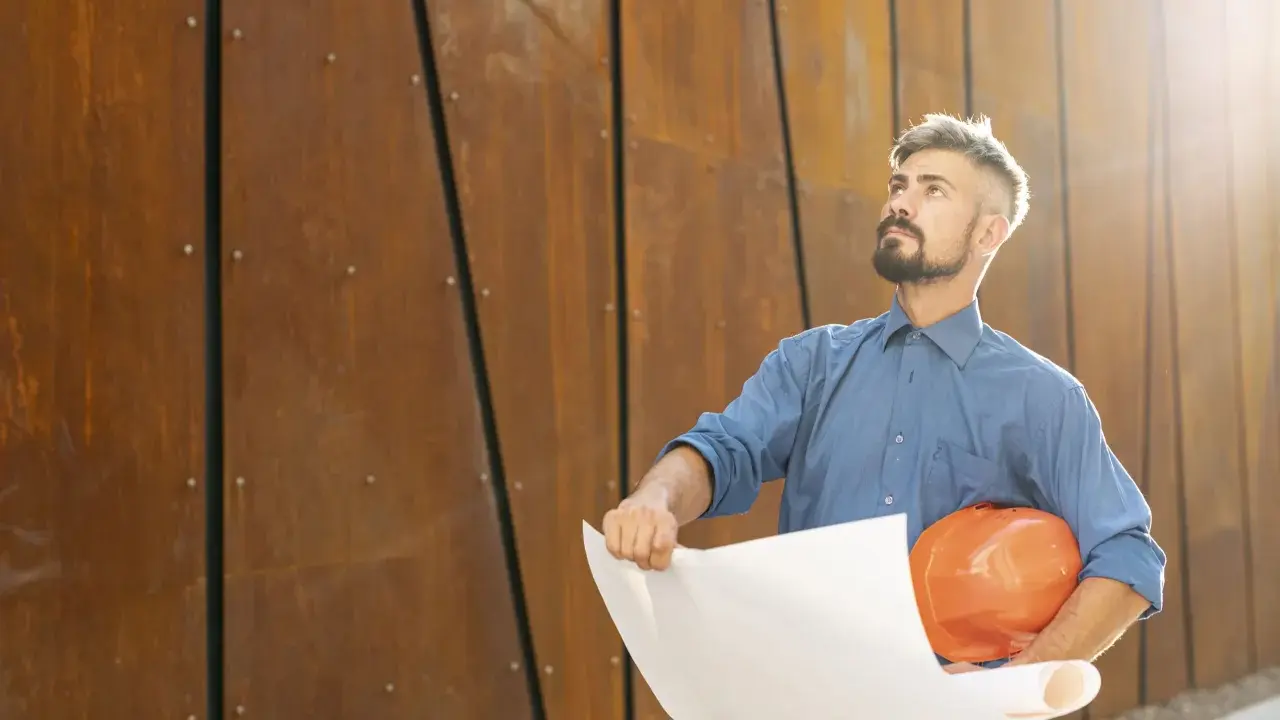 Un director de obra y director de ejecución, con casco naranja y planos, mira hacia arriba frente a una pared de metal oxidado.