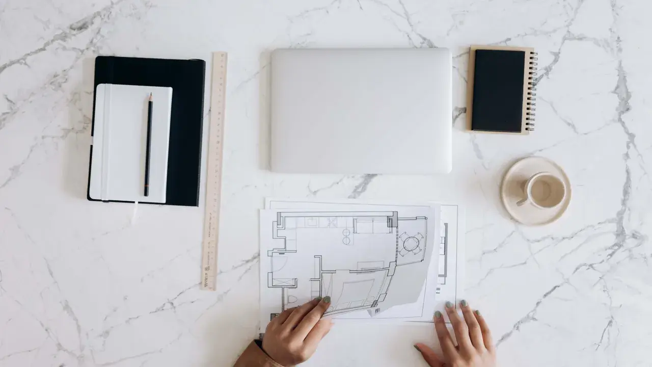 Manos revisando planos de reforma de vivienda para alquilar. Portátil, tazas y cuadernos listos para el proyecto.