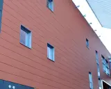 Fachada moderna con revestimiento de sate en tono terracota, ventanas rectangulares y una pasarela acristalada.