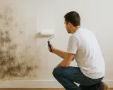 Hombre pintando pared con moho. La pintura caduca sin abrir, pero el problema de humedad persiste.