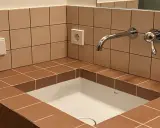 Lavabo empotrado con azulejos marrones y una **junta corrida** que delimita el espacio. Grifo cromado y enchufe visible.