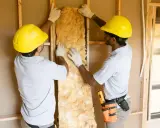 Dos obreros instalan aislamiento en una pared, eligiendo materiales con baja conductividad para mejorar la eficiencia energética.