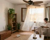 Mujer leyendo en sillón, con ventilador de techo y cortinas movidas por la brisa. Consejos aire acondicionado: ventilar y usar ventilador para refrescar.