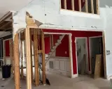 Interior en proceso de demolición, mostrando la estructura de madera expuesta y el cableado. Se observa una escalera roja y paredes parcialmente derribadas, como si se estuviera planeando como tirar una pared de casa.