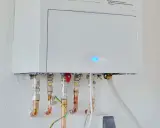 Calentador atmosférico blanco montado en pared con tuberías de cobre y conexiones. Una luz azul parpadea en la unidad, indicando que se puede instalar un calentador atmosférico en el interior.