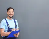 Técnico sonriente con mono azul y camiseta clara, revisando notas para el mantenimiento del aire acondicionado.