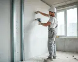 Obrero con casco instala tabiques de pladur, atornillando placas de yeso a una estructura metálica.