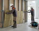 Dos obreros instalan aislamiento de lana de roca en una pared interior. Se ven herramientas y una caja de Rockwool.