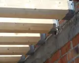 Detalle de la construcción: se están colocando vigas de madera sobre soportes metálicos anclados a una pared de ladrillo y hormigón.