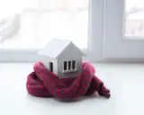 Casa de juguete envuelta en bufanda, simbolizando el aislamiento térmico y la baja conductividad para mantener el calor.