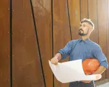 Un director de obra y director de ejecución, con casco naranja y planos, mira hacia arriba frente a una pared de metal oxidado.