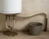 Un termo pierde agua por arriba, mojando el suelo y un cubo metálico espera bajo la tubería.