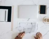 Manos revisando planos de reforma de vivienda para alquilar. Portátil, tazas y cuadernos listos para el proyecto.