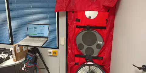 Equipo para blower door test en puerta, con laptop mostrando datos y medidor de presión.