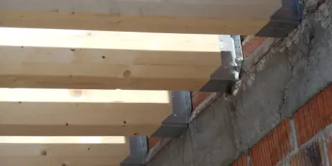 Detalle de la construcción: se están colocando vigas de madera sobre soportes metálicos anclados a una pared de ladrillo y hormigón.