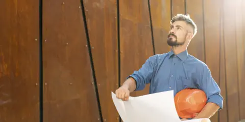 Un director de obra y director de ejecución, con casco naranja y planos, mira hacia arriba frente a una pared de metal oxidado.