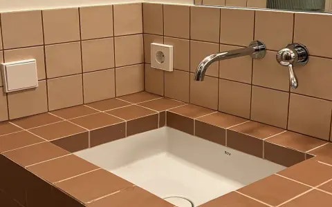Lavabo empotrado con azulejos marrones y una **junta corrida** que delimita el espacio. Grifo cromado y enchufe visible.