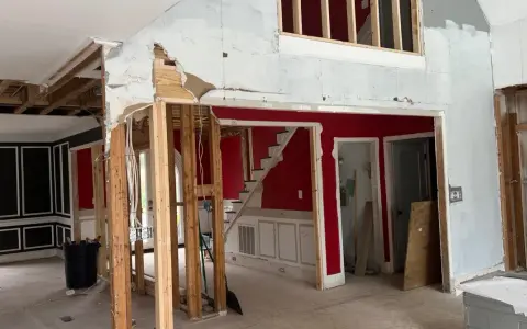 Interior en proceso de demolición, mostrando la estructura de madera expuesta y el cableado. Se observa una escalera roja y paredes parcialmente derribadas, como si se estuviera planeando como tirar una pared de casa.