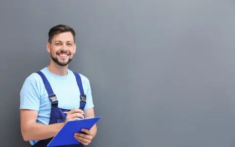 Técnico sonriente con mono azul y camiseta clara, revisando notas para el mantenimiento del aire acondicionado.