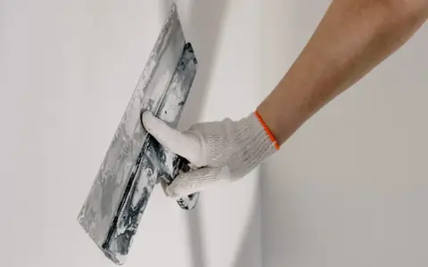 Mano enguantada aplicando yeso a una pared, parte del proceso de como alicatar una pared exterior.