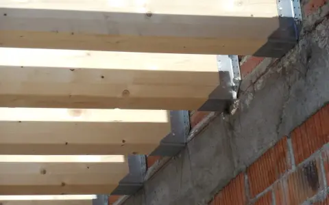 Detalle de la construcción: se están colocando vigas de madera sobre soportes metálicos anclados a una pared de ladrillo y hormigón.
