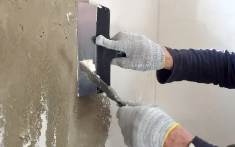 Manos enguantadas aplicando mortero para reparar pared.