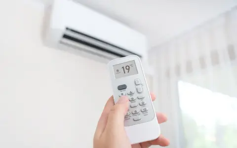 Mano sujetando control remoto de aire acondicionado, ajustando temperatura a 19°C. ¿Es mejor apagar el aire acondicionado o dejarlo encendido?