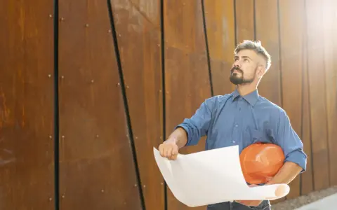 Un director de obra y director de ejecución, con casco naranja y planos, mira hacia arriba frente a una pared de metal oxidado.