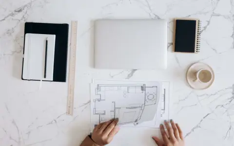 Manos revisando planos de reforma de vivienda para alquilar. Portátil, tazas y cuadernos listos para el proyecto.