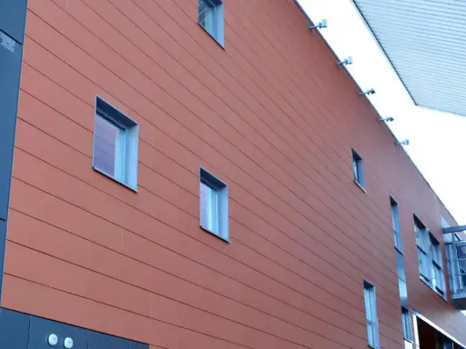 Fachada moderna con revestimiento de sate en tono terracota, ventanas rectangulares y una pasarela acristalada.