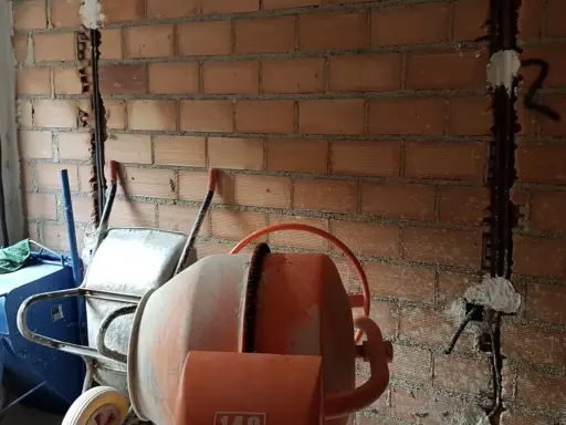 Preparativos para como tirar tabiques de ladrillo: carretilla, hormigonera y pared con ranuras para cables.