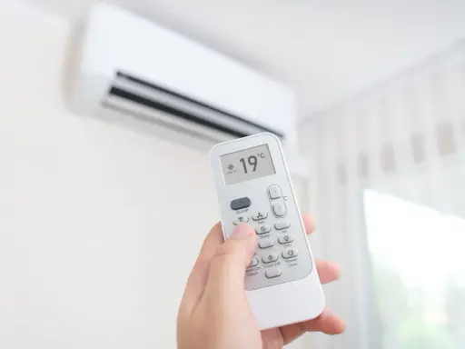 Mano sujetando control remoto de aire acondicionado, ajustando temperatura a 19°C. ¿Es mejor apagar el aire acondicionado o dejarlo encendido?