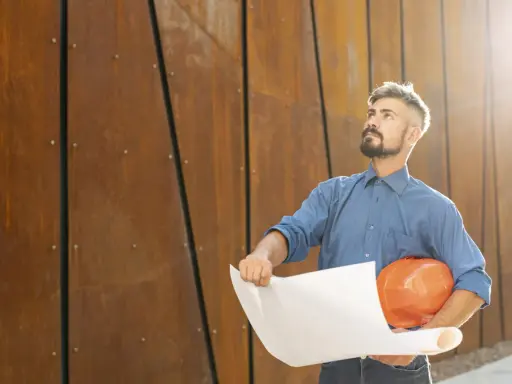 Un director de obra y director de ejecución, con casco naranja y planos, mira hacia arriba frente a una pared de metal oxidado.