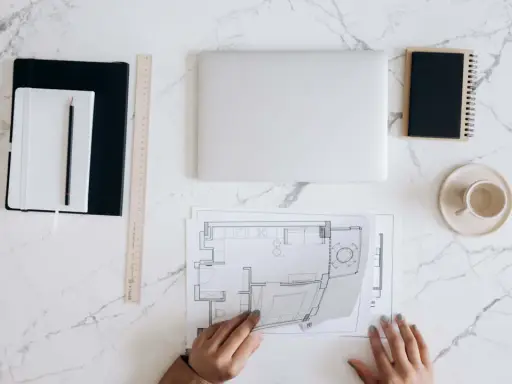 Manos revisando planos de reforma de vivienda para alquilar. Portátil, tazas y cuadernos listos para el proyecto.