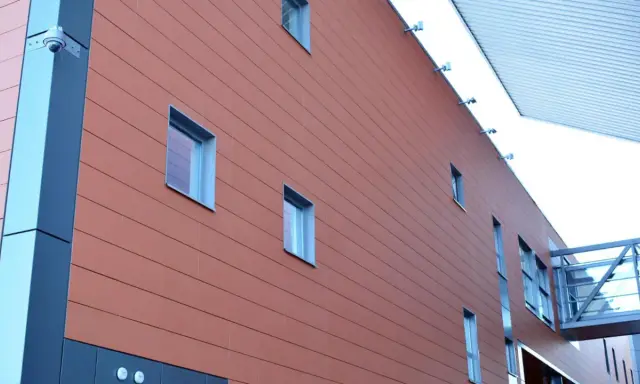 Fachada moderna con revestimiento de sate en tono terracota, ventanas rectangulares y una pasarela acristalada.