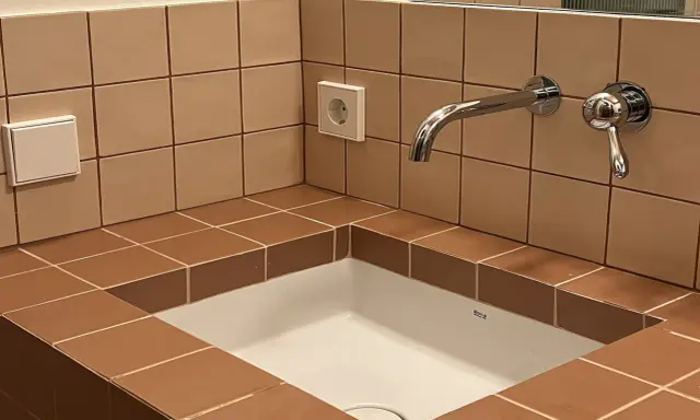 Lavabo empotrado con azulejos marrones y una **junta corrida** que delimita el espacio. Grifo cromado y enchufe visible.