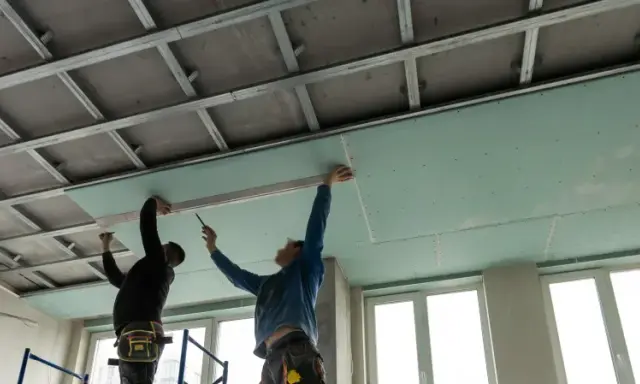 Dos trabajadores instalan placas de yeso para como aislar un techo por el interior. Se ve la estructura metálica del falso techo.