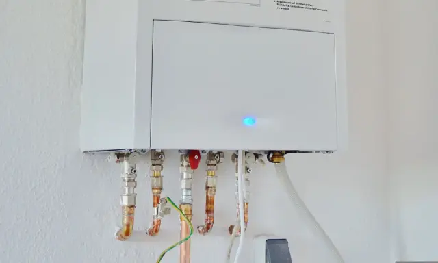 Calentador atmosférico blanco montado en pared con tuberías de cobre y conexiones. Una luz azul parpadea en la unidad, indicando que se puede instalar un calentador atmosférico en el interior.