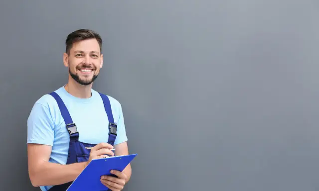 Técnico sonriente con mono azul y camiseta clara, revisando notas para el mantenimiento del aire acondicionado.
