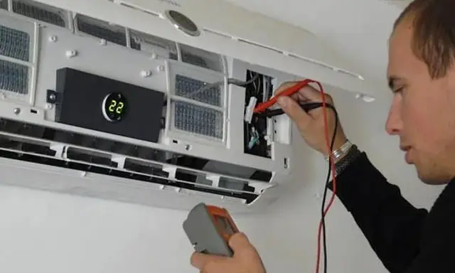 Técnico revisa unidad interior de aire acondicionado con multímetro, cumpliendo la normativa de instalación.