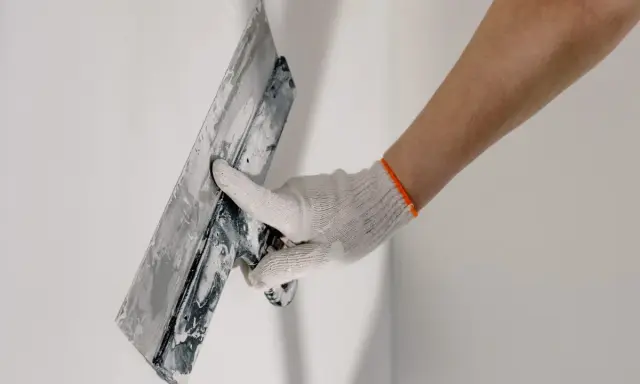 Mano enguantada aplicando yeso a una pared, parte del proceso de como alicatar una pared exterior.