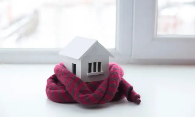 Casa de juguete envuelta en bufanda, simbolizando el aislamiento térmico y la baja conductividad para mantener el calor.