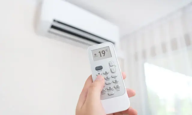 Mano sujetando control remoto de aire acondicionado, ajustando temperatura a 19°C. ¿Es mejor apagar el aire acondicionado o dejarlo encendido?