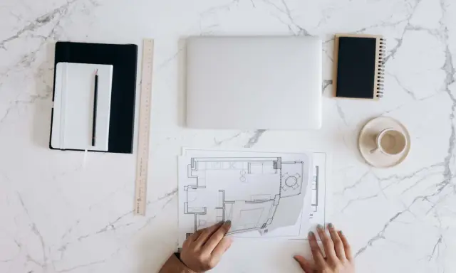 Manos revisando planos de reforma de vivienda para alquilar. Portátil, tazas y cuadernos listos para el proyecto.