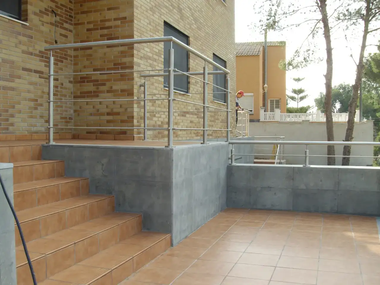 Escaleras y terraza de baldosas, con barandillas de acero inoxidable. Un hombre con gorra roja supervisa la obra para hacer una terraza.